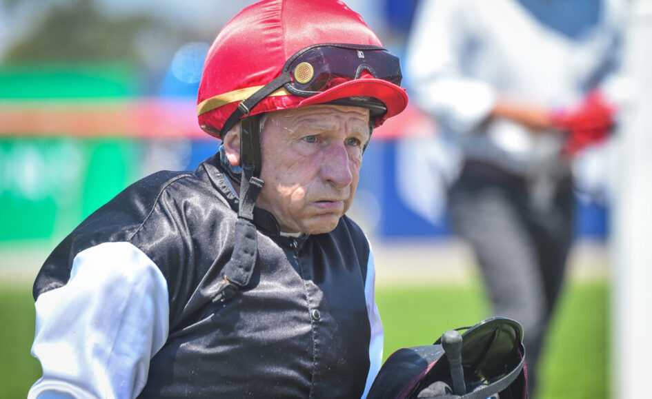 Older jockey wearing a red helmet and black riding vest, holding a racing saddle, outdoors on a sunny day