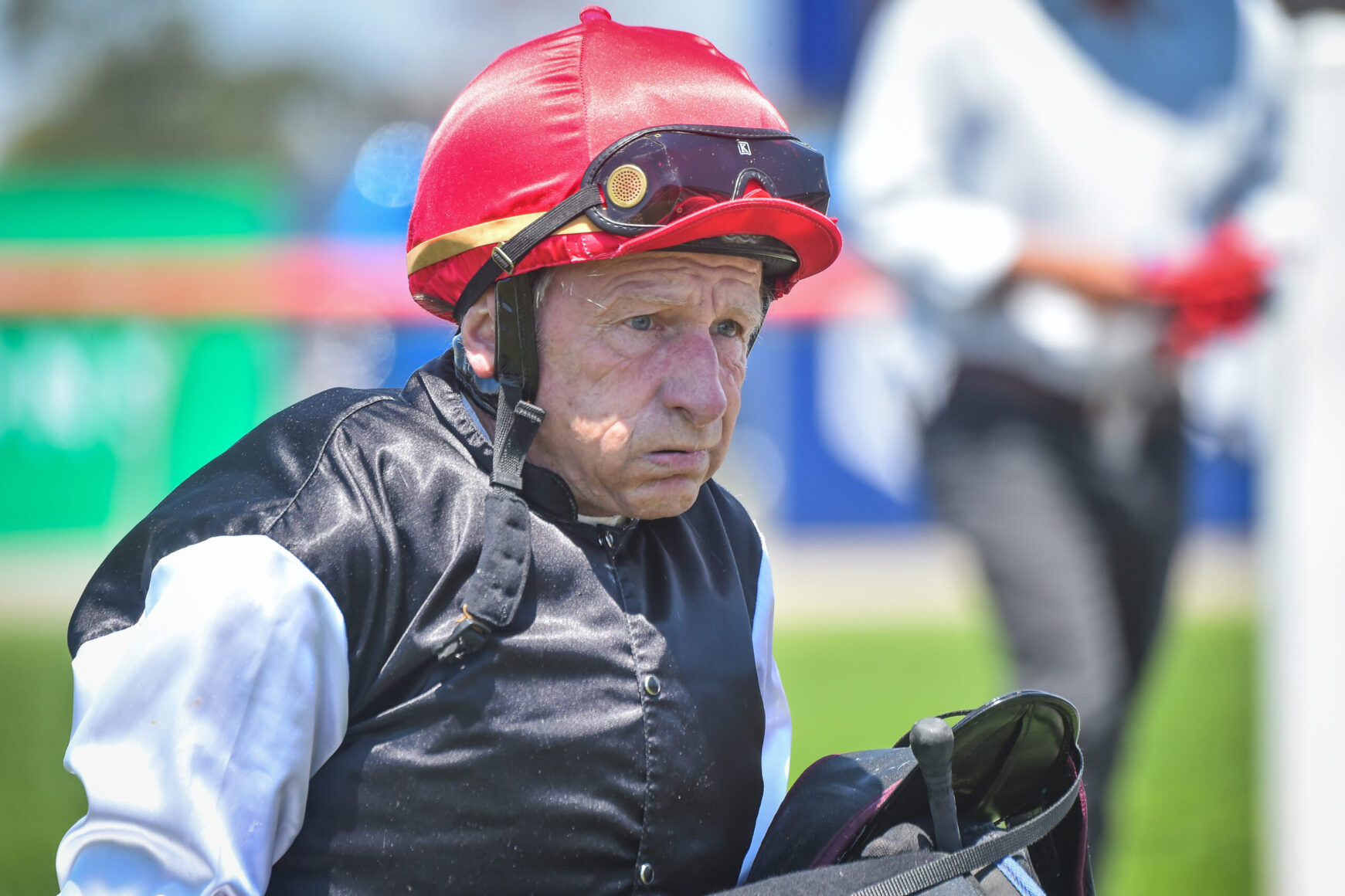 Older jockey wearing a red helmet and black riding vest, holding a racing saddle, outdoors on a sunny day