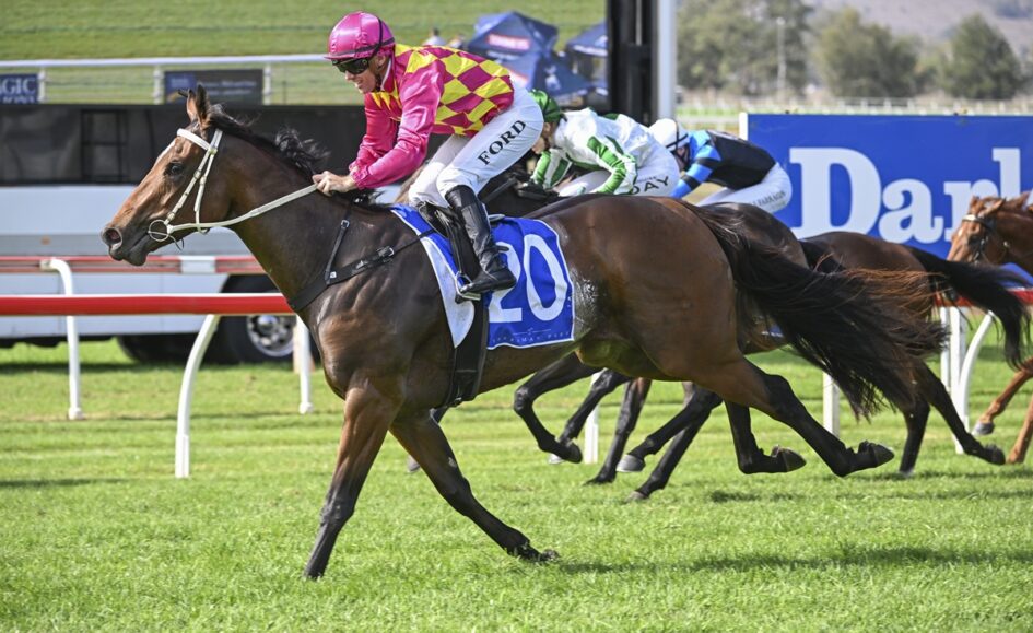 A jockey in pink and yellow silks rides a brown horse, leading a race on a grassy track with other horses close behind.