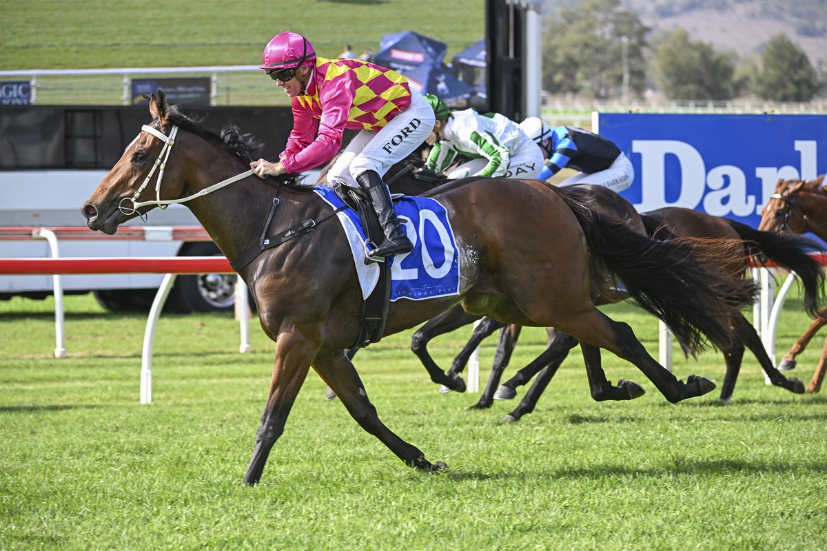 A jockey in pink and yellow silks rides a brown horse, leading a race on a grassy track with other horses close behind.