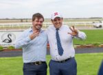 Two men in dress shirts posing outdoors at a racecourse, flashing victory signs and smiling at the camera.