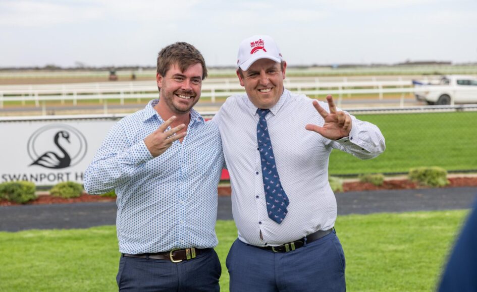 Two men in dress shirts posing outdoors at a racecourse, flashing victory signs and smiling at the camera.