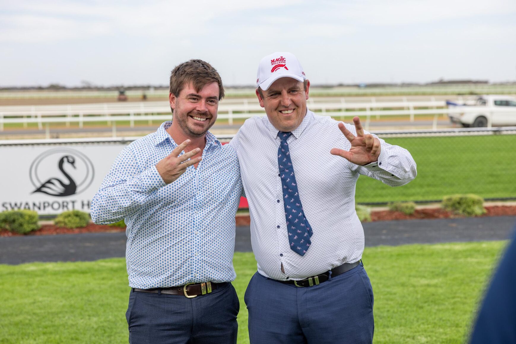 Two men in dress shirts posing outdoors at a racecourse, flashing victory signs and smiling at the camera.