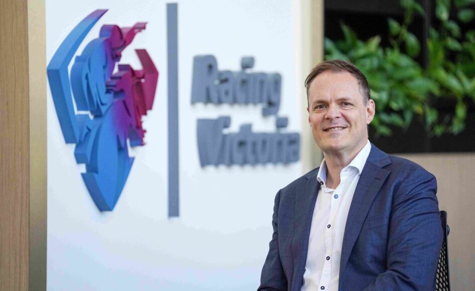 Smiling man in a blue suit seated in an office with a colorful 3D logo on the wall behind him.