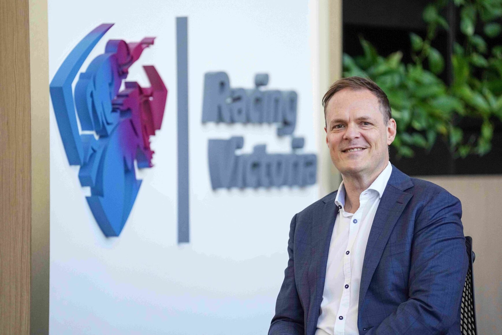 Smiling man in a blue suit seated in an office with a colorful 3D logo on the wall behind him.