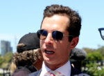 Man in a navy suit and pink striped tie wearing Ray-Ban sunglasses at an outdoor event.