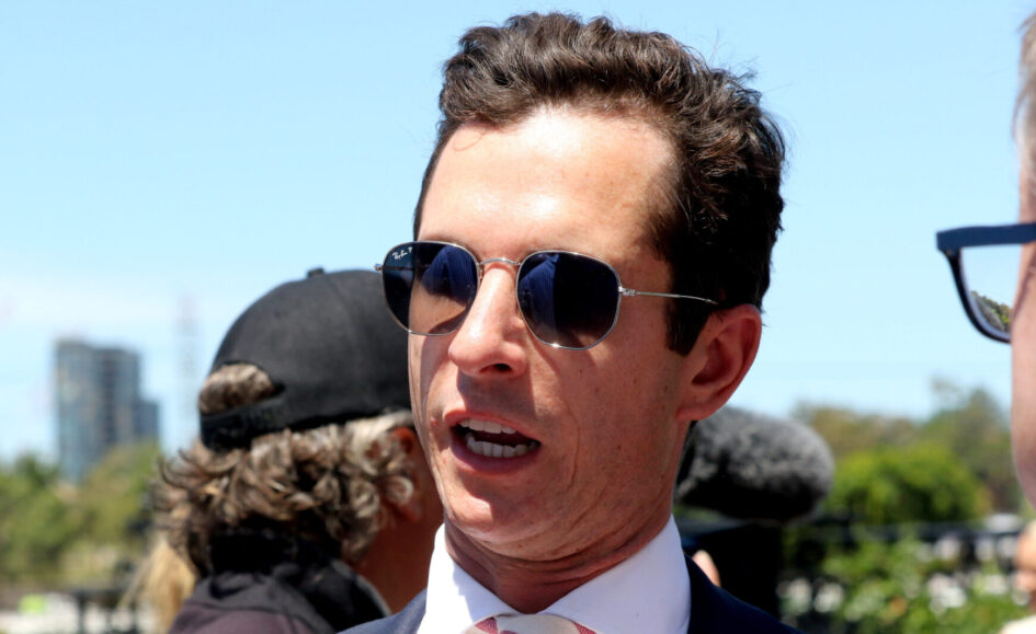 Man in a navy suit and pink striped tie wearing Ray-Ban sunglasses at an outdoor event.