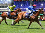 Two jockeys race their brown horses to the finish line on a sunny track, green grass in the foreground and purple banners in the background.