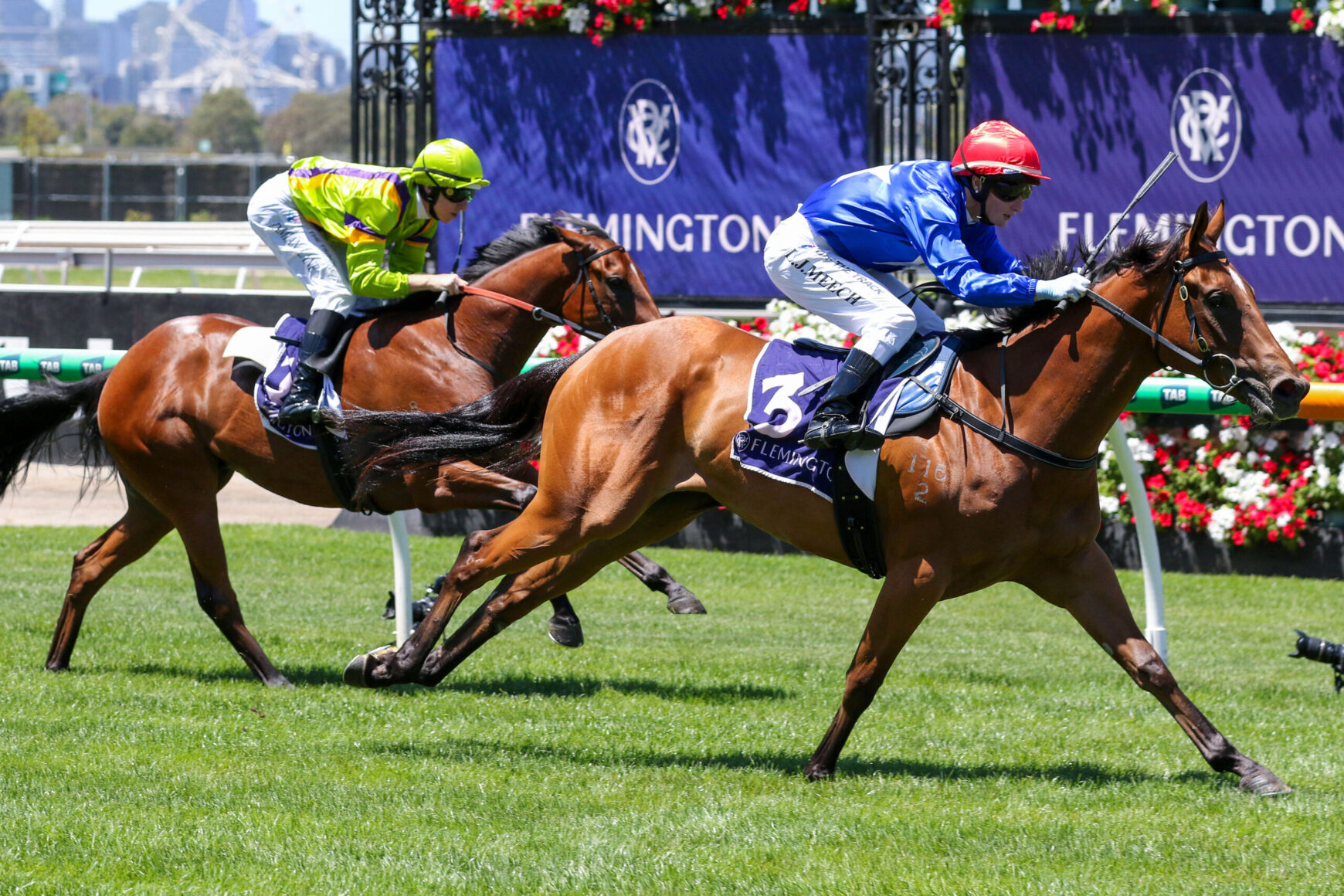 Two jockeys race their brown horses to the finish line on a sunny track, green grass in the foreground and purple banners in the background.