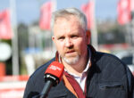 Middle-aged man being interviewed outdoors, speaking into a red Racing.com microphone, wearing a dark quilted jacket.
