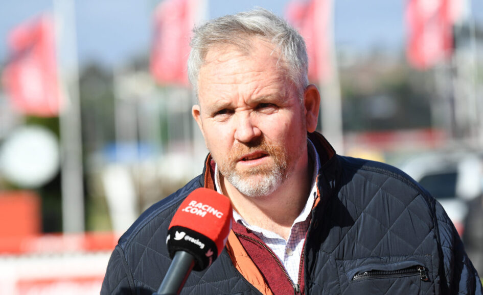 Middle-aged man being interviewed outdoors, speaking into a red Racing.com microphone, wearing a dark quilted jacket.