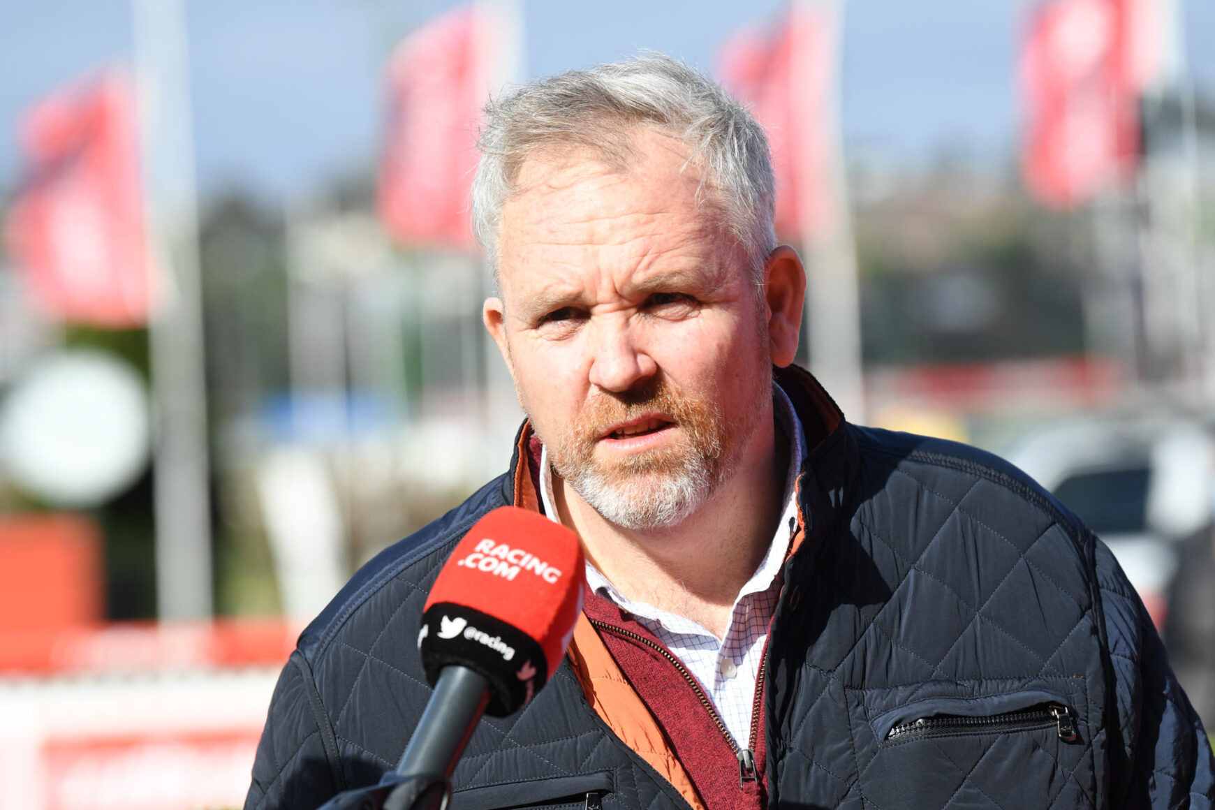 Middle-aged man being interviewed outdoors, speaking into a red Racing.com microphone, wearing a dark quilted jacket.