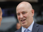Bald man in a navy suit and striped tie speaks during an outdoor conversation.