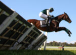 Brown horse and rider in green and white gear leap over a wooden obstacle against a clear blue sky.