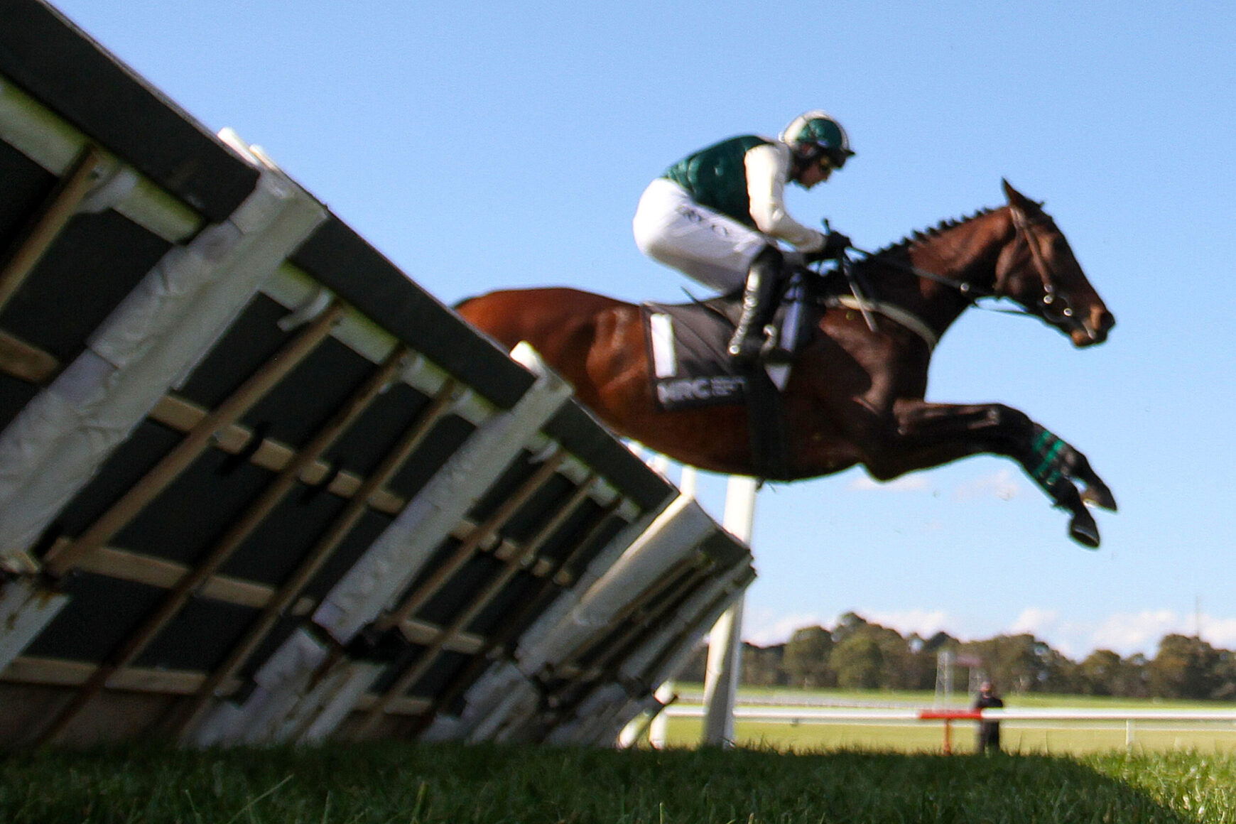 Brown horse and rider in green and white gear leap over a wooden obstacle against a clear blue sky.