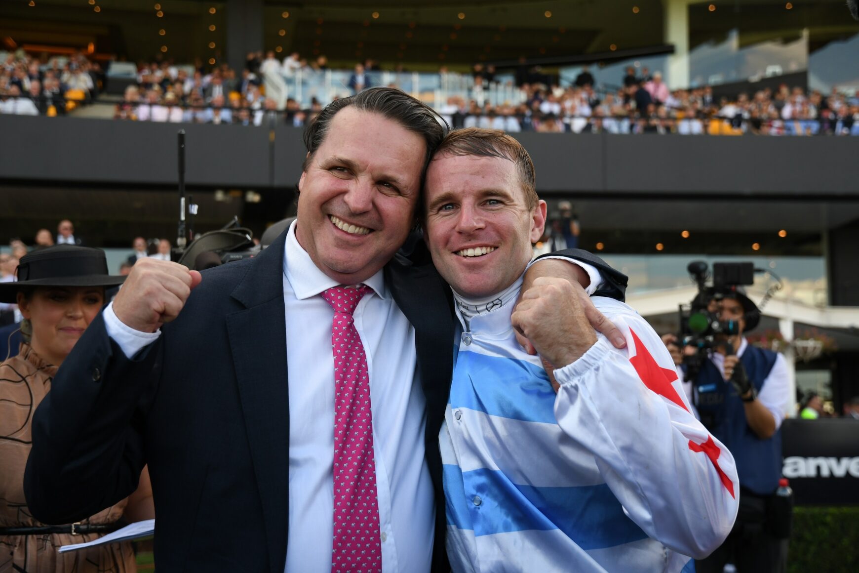 Two men pose and smile with arms around each other at a crowded event.