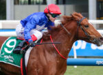Jockey in blue silks and red helmet riding a chestnut horse during a race, green saddlecloth visible on the hindquarters.