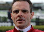 Close-up of a man in a red racing suit outdoors, looking serious with a furrowed brow.