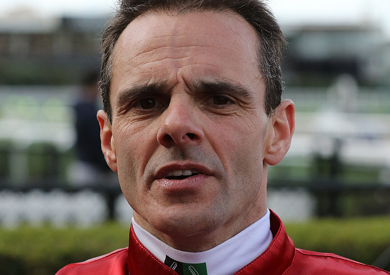Close-up of a man in a red racing suit outdoors, looking serious with a furrowed brow.