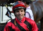Jockey in red-and-black striped racing silks and a red helmet smiling at the camera