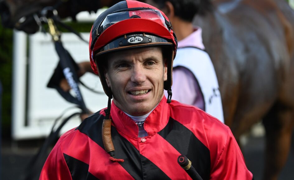 Jockey in red-and-black striped racing silks and a red helmet smiling at the camera