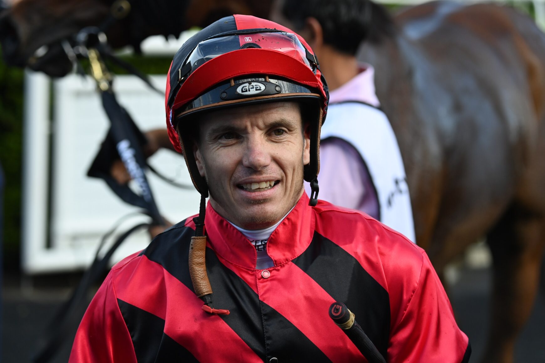 Jockey in red-and-black striped racing silks and a red helmet smiling at the camera