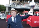 Man in a navy suit and straw hat raises a large silver trophy at an outdoor event, with red barriers and spectators in the background.”,