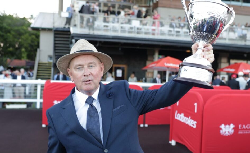 Man in a navy suit and straw hat raises a large silver trophy at an outdoor event, with red barriers and spectators in the background.”,