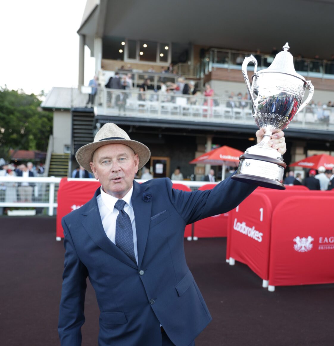 Man in a navy suit and straw hat raises a large silver trophy at an outdoor event, with red barriers and spectators in the background.”,