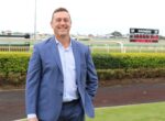 Man in a blue suit smiling at the camera at a racecourse with a leaderboard in the background