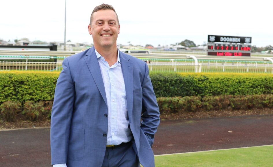 Man in a blue suit smiling at the camera at a racecourse with a leaderboard in the background