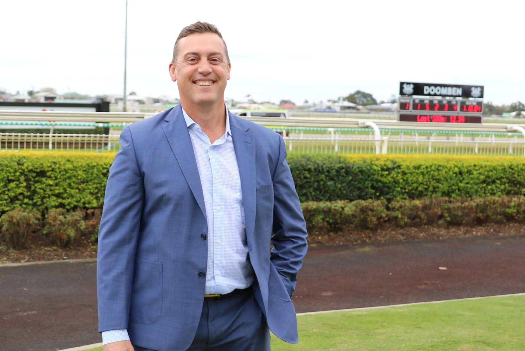 Man in a blue suit smiling at the camera at a racecourse with a leaderboard in the background
