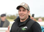 Smiling man wearing a cap and black jacket outdoors, giving a thumbs-up gesture.