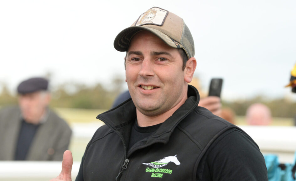Smiling man wearing a cap and black jacket outdoors, giving a thumbs-up gesture.