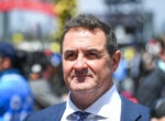 Portrait of a middle-aged man in a blue suit and patterned tie, outdoors at a crowd-filled event, smiling slightly.