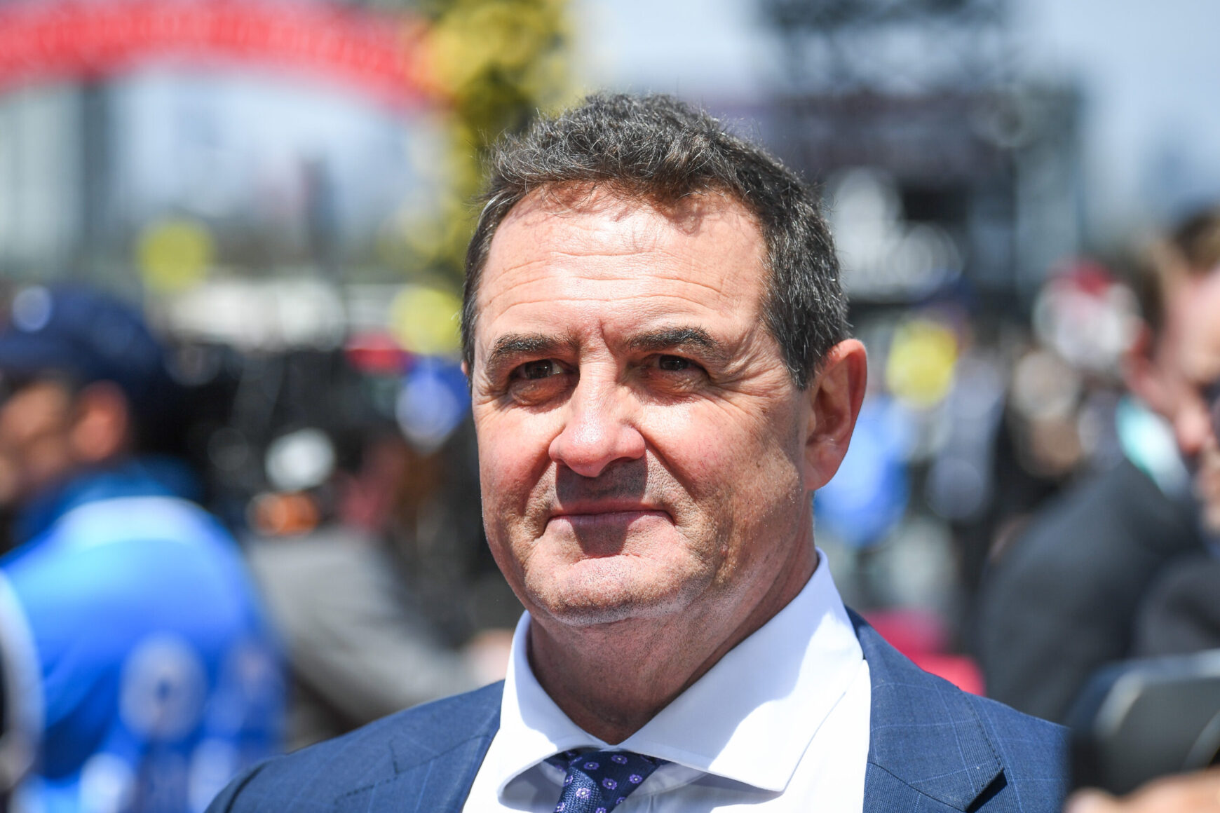 Portrait of a middle-aged man in a blue suit and patterned tie, outdoors at a crowd-filled event, smiling slightly.