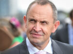 Close-up of a middle-aged man in a gray suit and pink tie, outdoors, with a slight smile on his face.